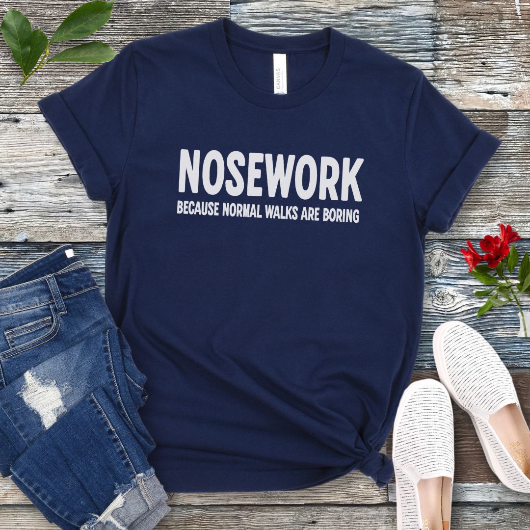 Navy blue K9 Nose Work t-shirt with 'NOSEWORK BECAUSE NORMAL WALKS ARE BORING' text on a wooden surface with jeans and shoes.