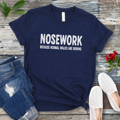 Navy blue K9 Nose Work t-shirt with 'NOSEWORK BECAUSE NORMAL WALKS ARE BORING' text on a wooden surface with jeans and shoes.