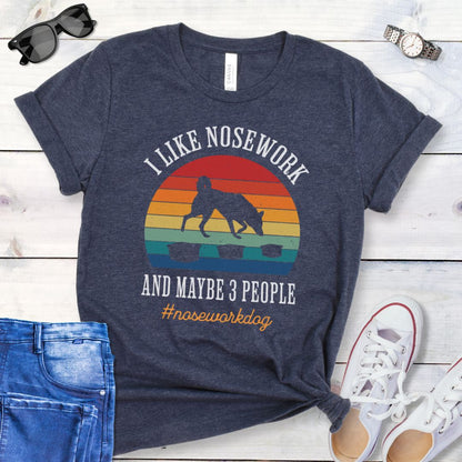 Heather navy nosework t-shirt with sniffing dog graphic and "I Like Nosework and Maybe 3 People" text on a wooden surface with jeans and shoes.