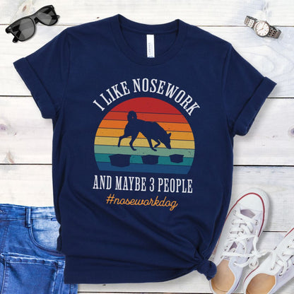 Navy blue nosework t-shirt with sniffing dog graphic and "I Like Nosework and Maybe 3 People" text on a wooden surface with jeans and shoes.