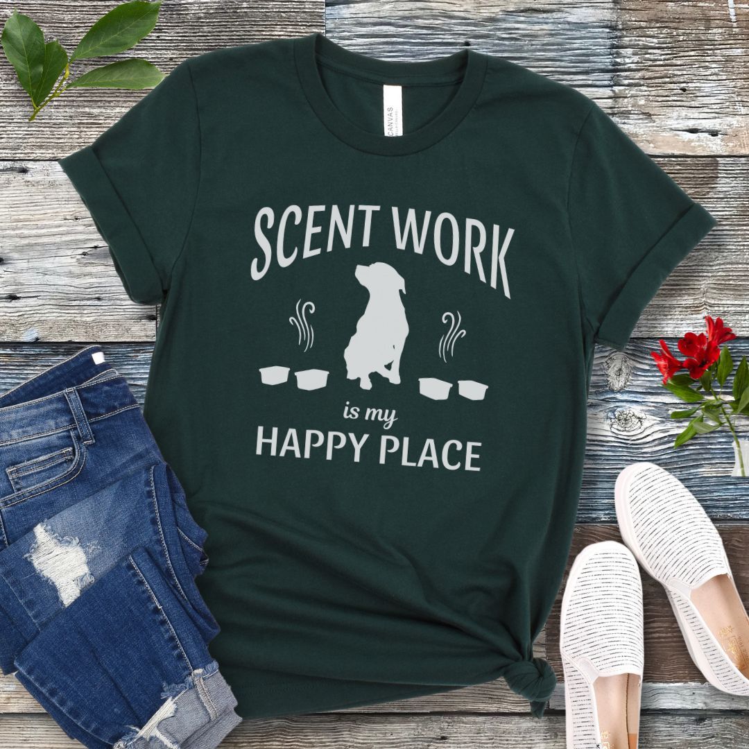 Dark green t-shirt with 'Scent Work is my Happy Place' text and dog silhouette on a wooden surface with jeans and shoes.