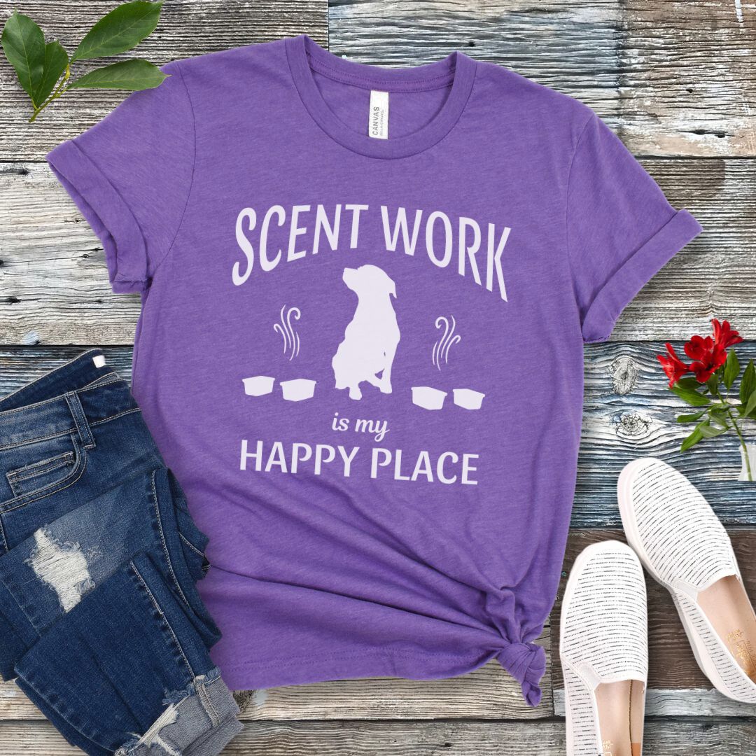 Purple t-shirt with 'Scent Work is my Happy Place' text and dog silhouette on a wooden surface with jeans and shoes.