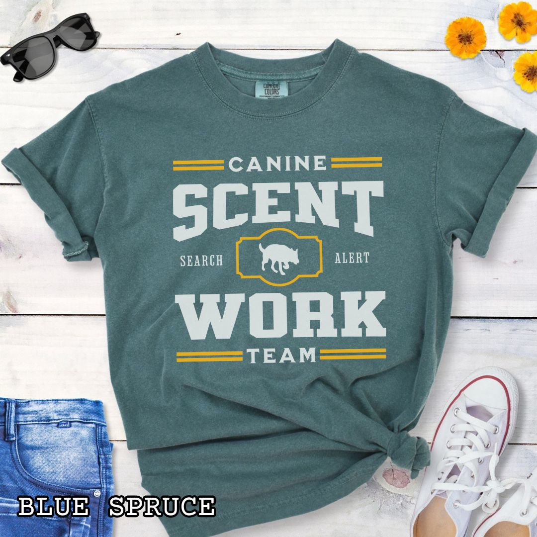 Green Comfort Colors t-shirt with 'Canine Scent Work Team' and custom breed silhouette, presented text on a wooden surface with shoes and jeans.