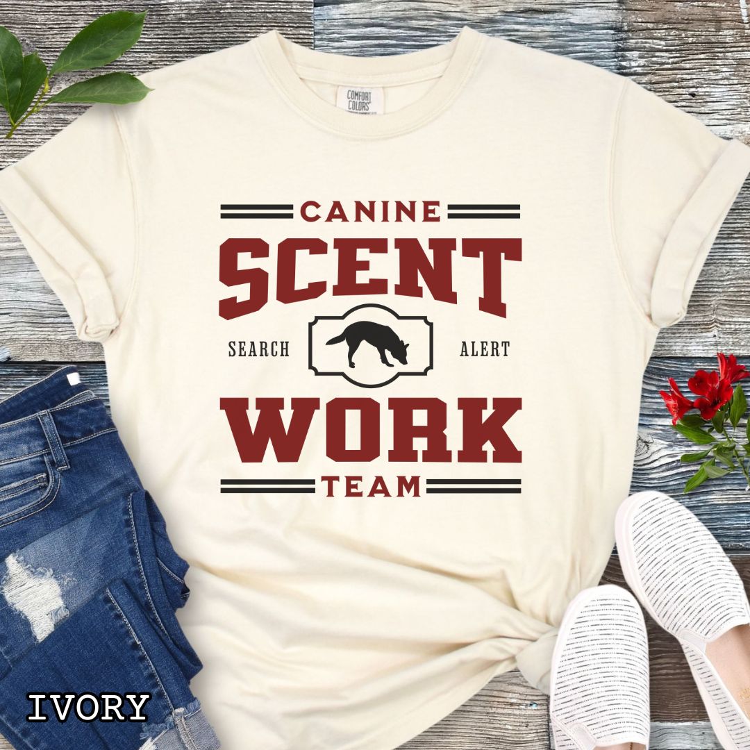 Beige Comfort Colors t-shirt with 'Canine Scent Work Team' and custom breed silhouette, presented text on a wooden surface with shoes and jeans.