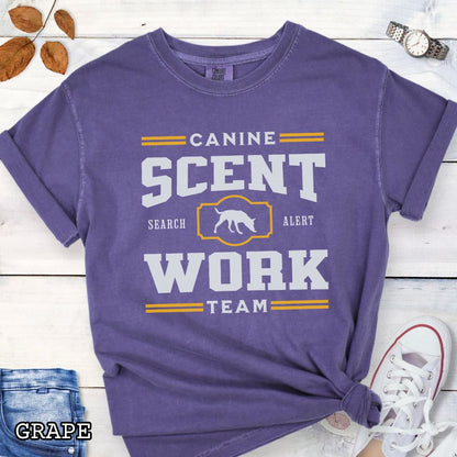 Purple Comfort Colors t-shirt with 'Canine Scent Work Team' and custom breed silhouette, presented text on a wooden surface with shoes and jeans.