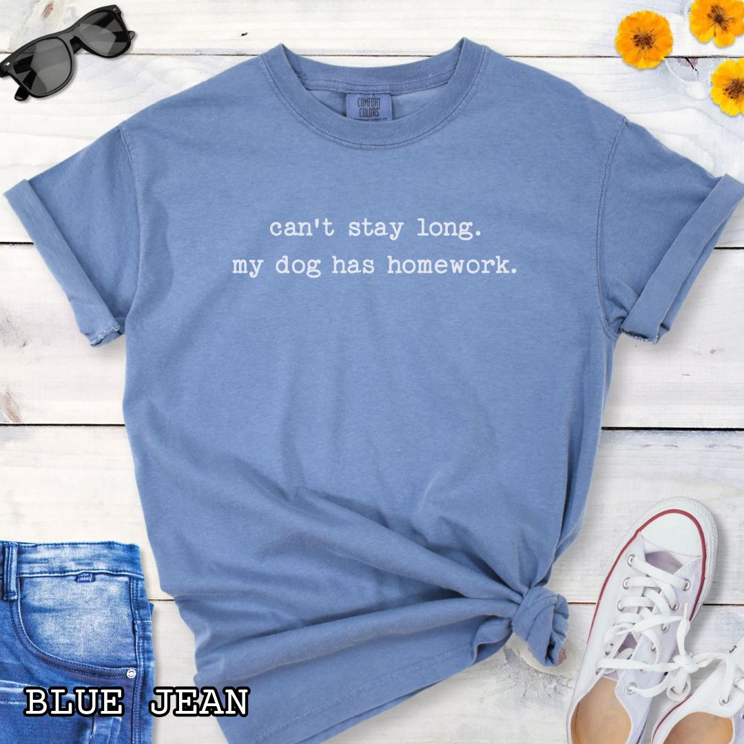 Blue Comfort Colors t-shirt with humorous text 'can't stay long. my dog has homework.' in minimalist typewriter lettering, shown on a wooden surface with sunglasses and jeans.