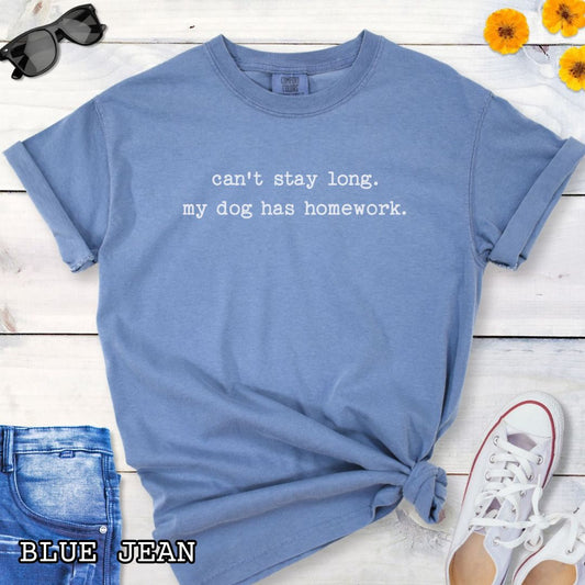Blue Comfort Colors t-shirt with humorous text 'can't stay long. my dog has homework.' in minimalist typewriter lettering, shown on a wooden surface with sunglasses and jeans.