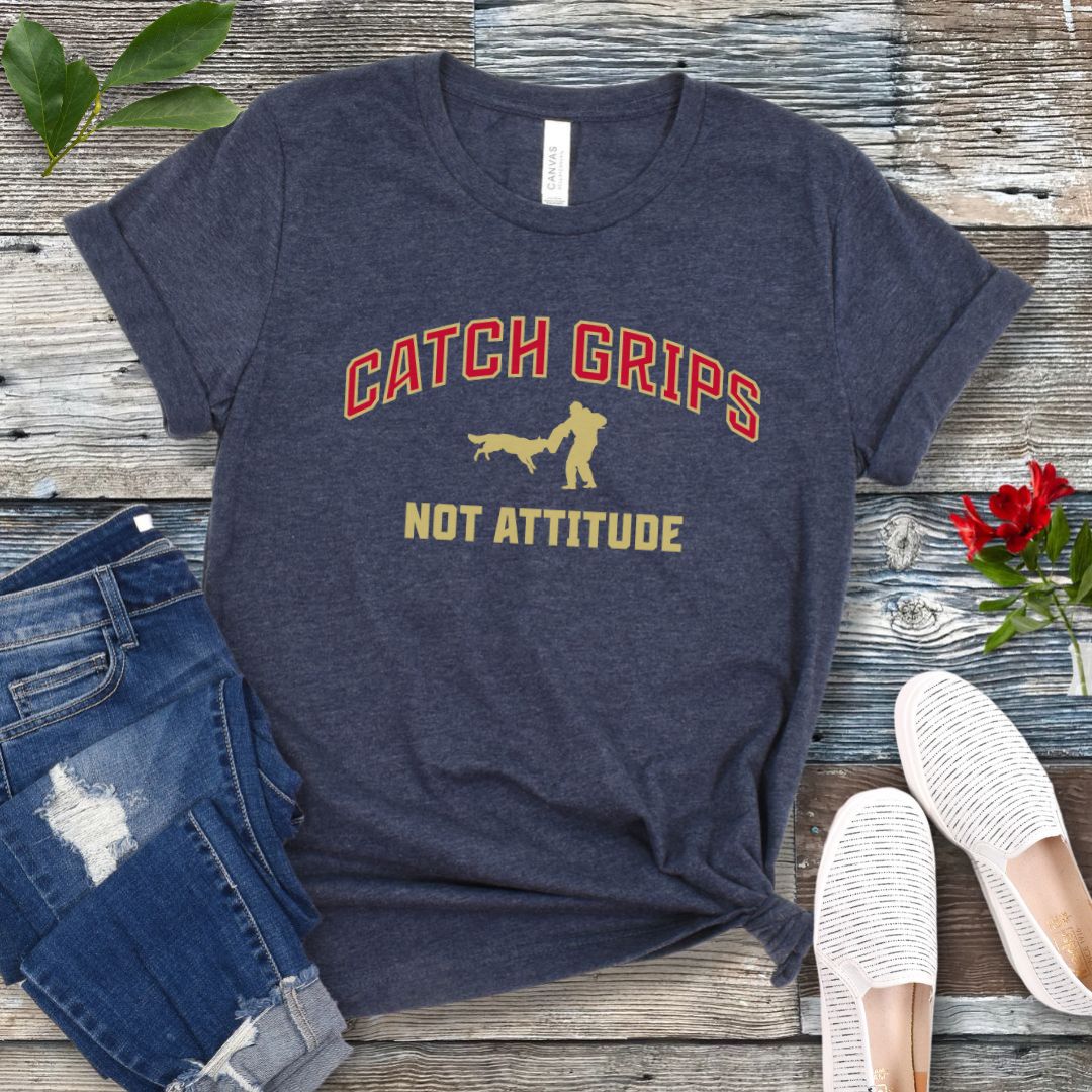 Heather navy t-shirt with 'Catch Grips Not Attitude' text and bitework graphic on a wooden surface with jeans and shoes.