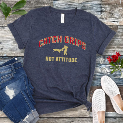 Heather navy t-shirt with 'Catch Grips Not Attitude' text and bitework graphic on a wooden surface with jeans and shoes.