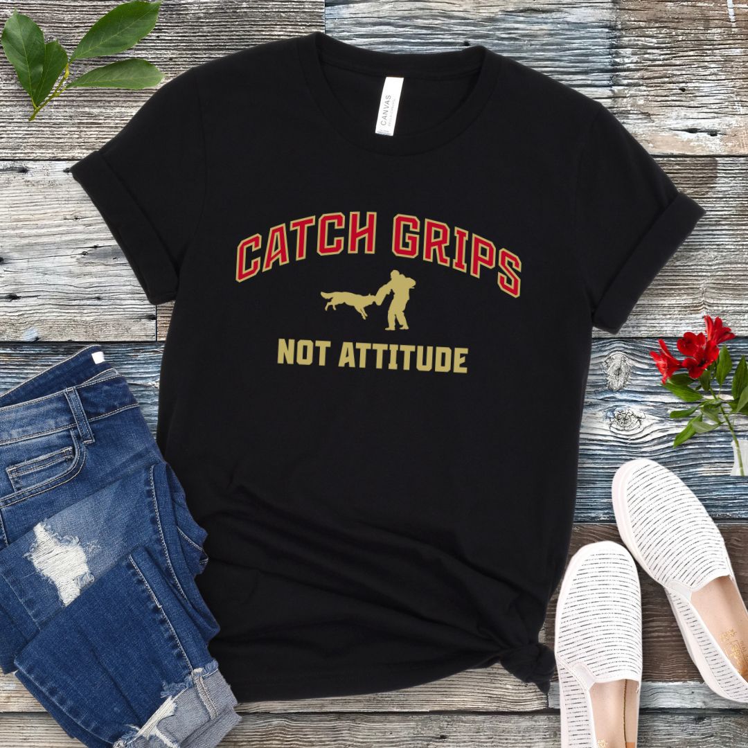 Black t-shirt with 'Catch Grips Not Attitude' text and bitework graphic on a wooden surface with jeans and shoes.