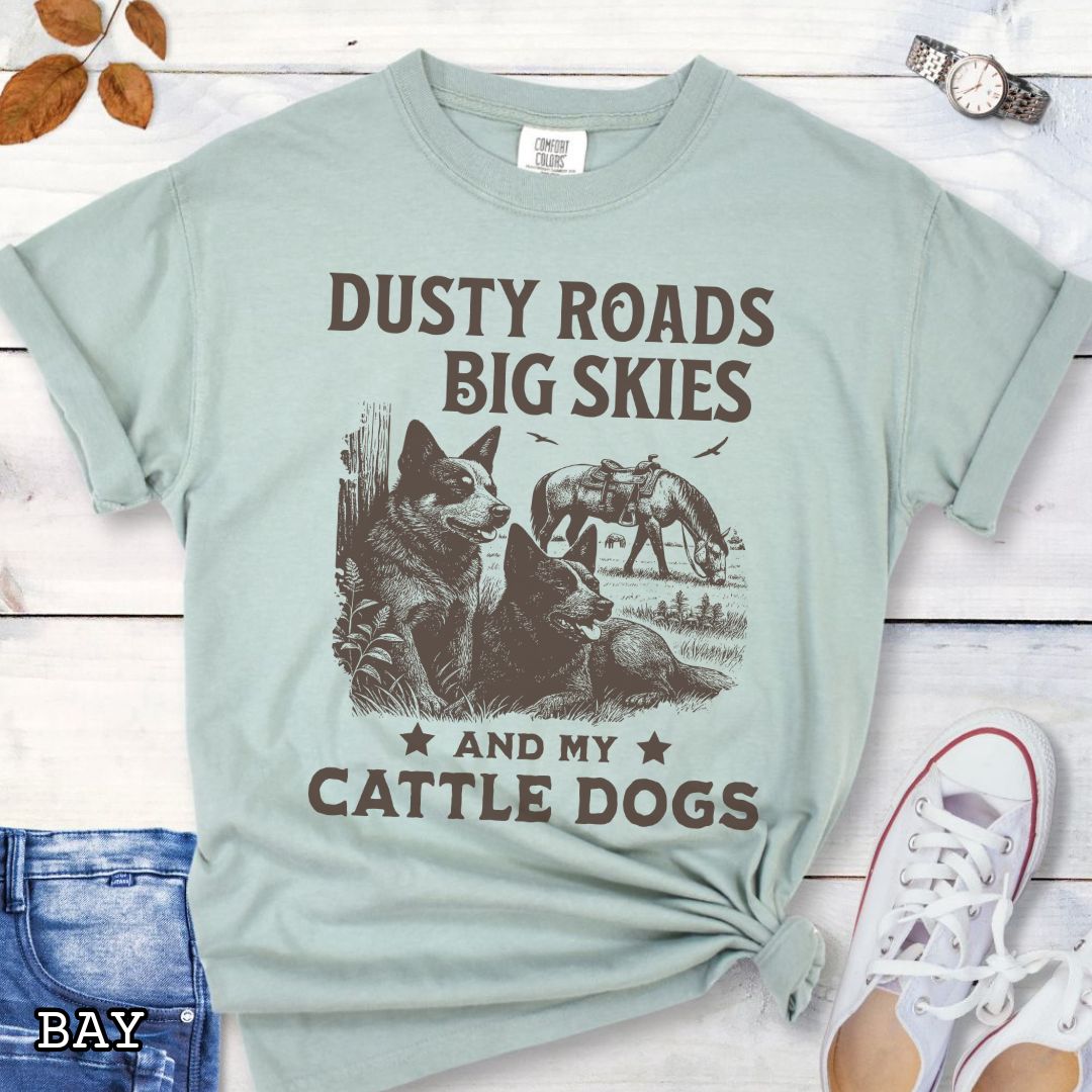 Light green Comfort Colors Cattle Dog t-shirt featuring a Western-style design with grazing horse, rustic fence, and Cattle Dog illustration; text reads “Dusty Roads, Big Skies, and My Cattle Dogs.”