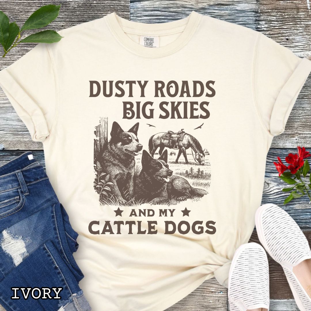 Ivory Comfort Colors Cattle Dog t-shirt featuring a Western-style design with grazing horse, rustic fence, and Cattle Dog illustration; text reads “Dusty Roads, Big Skies, and My Cattle Dogs.”