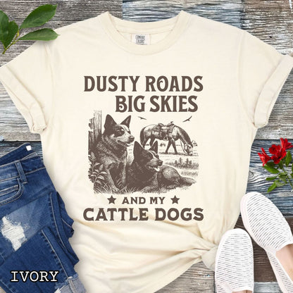 Ivory Comfort Colors Cattle Dog t-shirt featuring a Western-style design with grazing horse, rustic fence, and Cattle Dog illustration; text reads “Dusty Roads, Big Skies, and My Cattle Dogs.”