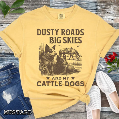 Mustard-yellow Comfort Colors Cattle Dog t-shirt featuring a Western-style design with grazing horse, rustic fence, and Cattle Dog illustration; text reads “Dusty Roads, Big Skies, and My Cattle Dogs.”