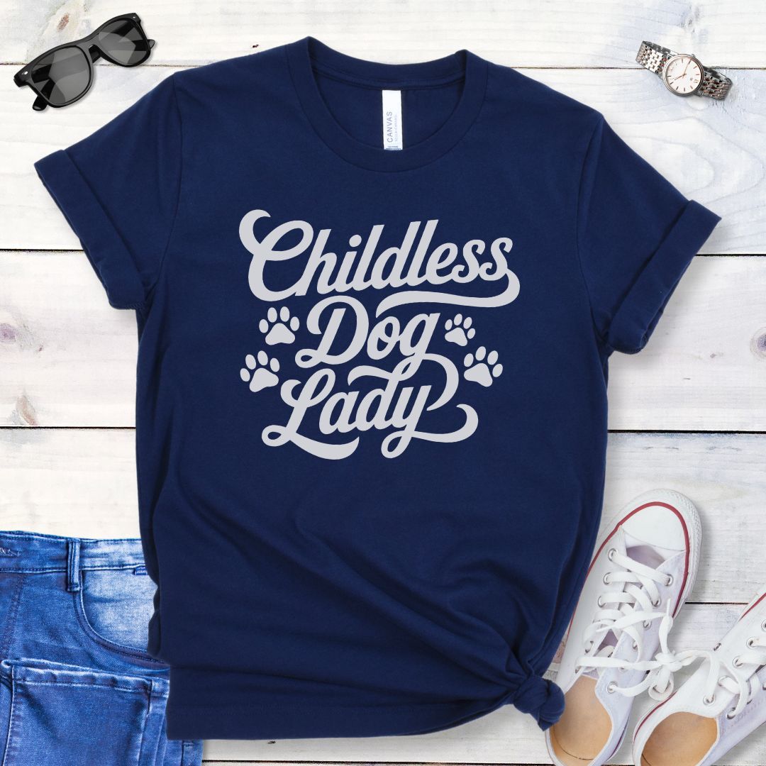 A navy blue dog lover t-shirt with the text 'Childless Dog Lady' in a script typography design, accented by paw prints. The shirt is displayed on a wooden surface, accompanied by a pair of blue jeans and white shoes.