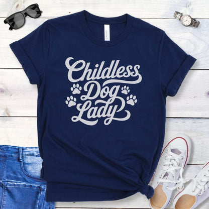 A navy blue dog lover t-shirt with the text 'Childless Dog Lady' in a script typography design, accented by paw prints. The shirt is displayed on a wooden surface, accompanied by a pair of blue jeans and white shoes.