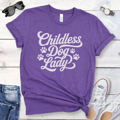 A purple dog lover t-shirt with the text 'Childless Dog Lady' in a script typography design, accented by paw prints. The shirt is displayed on a wooden surface, accompanied by a pair of blue jeans and white shoes.