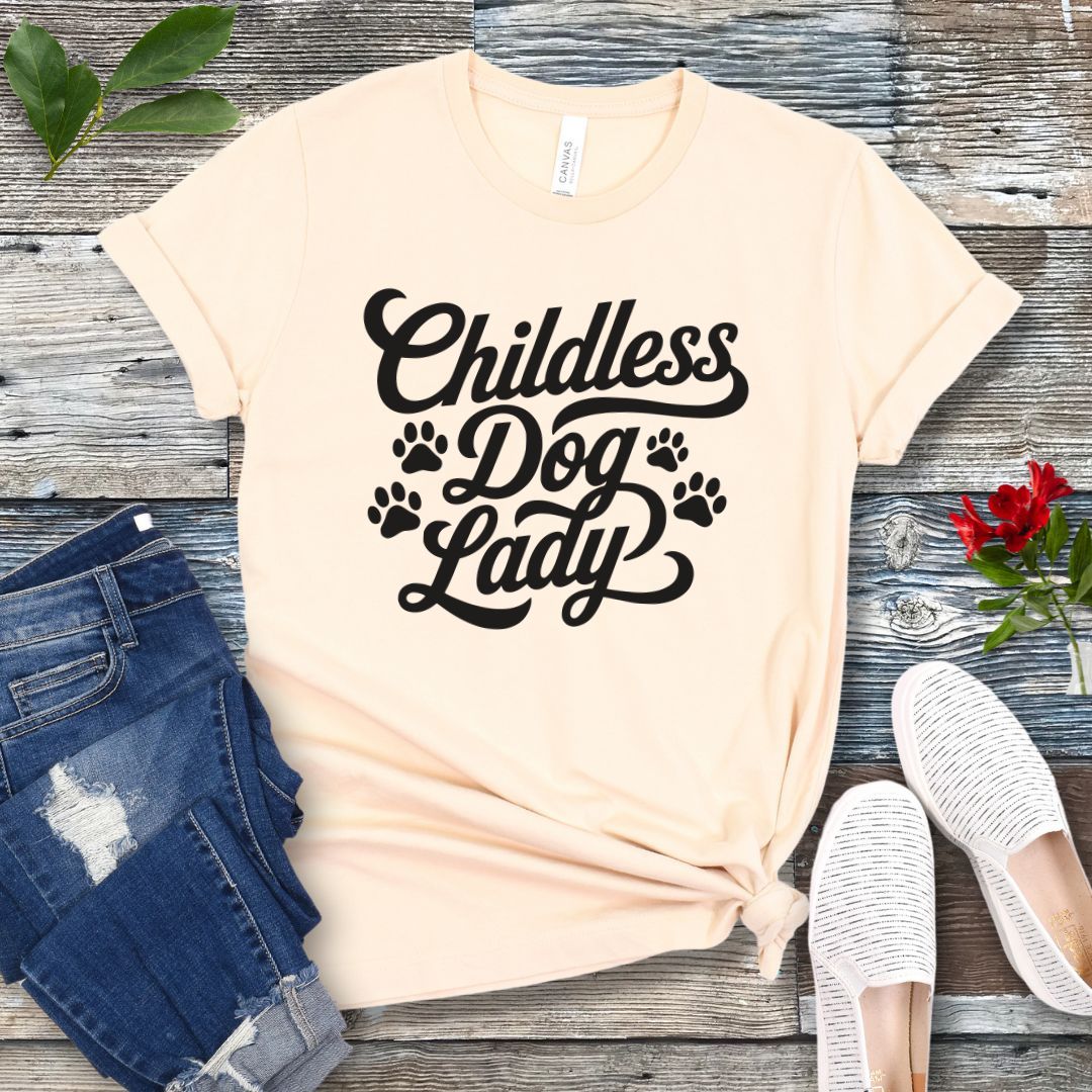 A beige colored dog lover t-shirt with the text 'Childless Dog Lady' in a script typography design, accented by paw prints. The shirt is displayed on a wooden surface, accompanied by a pair of blue jeans and white shoes.