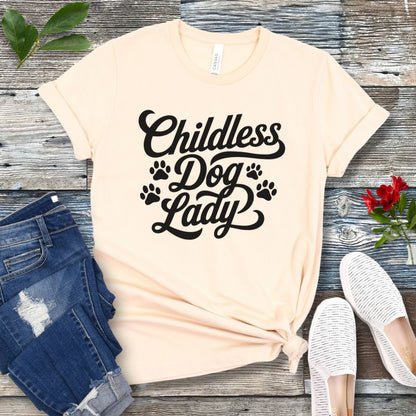A beige colored dog lover t-shirt with the text 'Childless Dog Lady' in a script typography design, accented by paw prints. The shirt is displayed on a wooden surface, accompanied by a pair of blue jeans and white shoes.