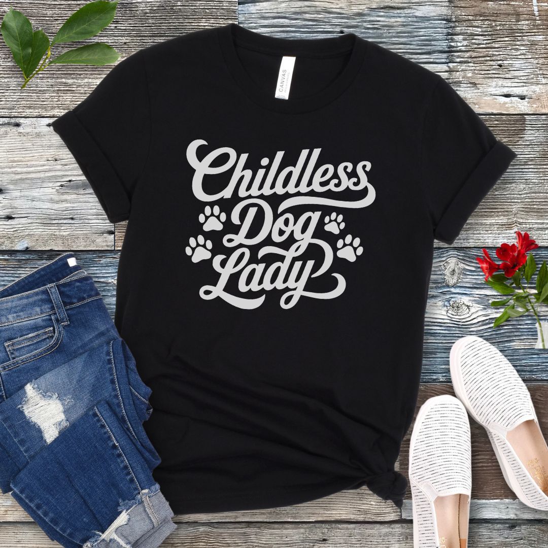 A black unisex dog lover t-shirt with the text 'Childless Dog Lady' in a script typography design, accented by paw prints. The shirt is displayed on a wooden surface, accompanied by a pair of blue jeans and white shoes.