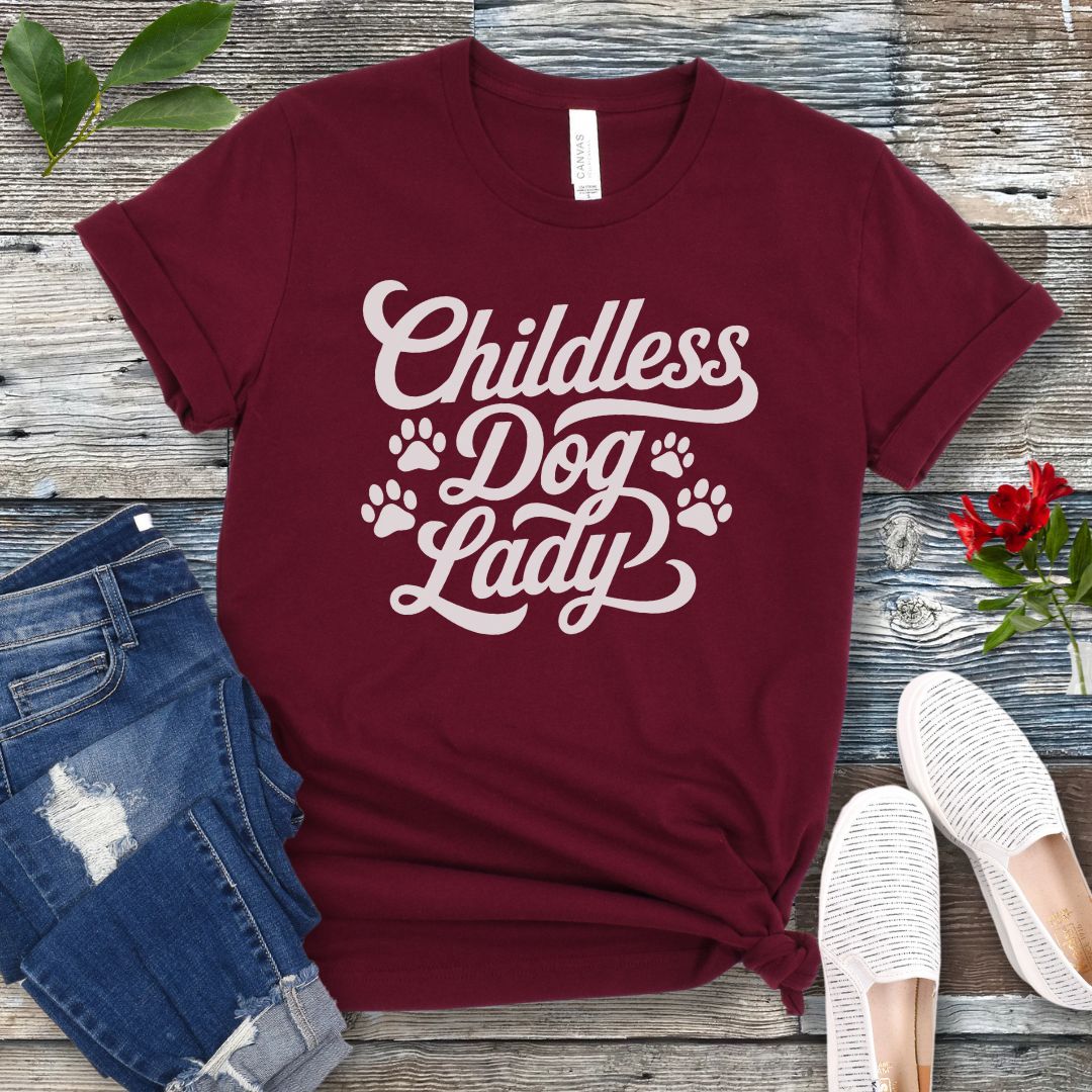 A maroon colored dog lover t-shirt with the text 'Childless Dog Lady' in a script typography design, accented by paw prints. The shirt is displayed on a wooden surface, accompanied by a pair of blue jeans and white shoes.