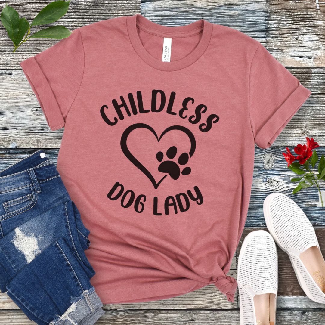 A mauve t-shirt dog dog lovers with the text 'CHILDLESS DOG LADY' in playful lettering and a heart and paw print graphic, displayed on a wooden surface with a pair of jeans and white shoes in the background.