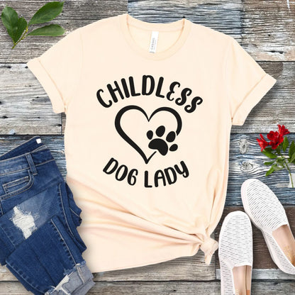 A beige t-shirt dog dog lovers with the text 'CHILDLESS DOG LADY' in playful lettering and a heart and paw print graphic, displayed on a wooden surface with a pair of jeans and white shoes in the background.