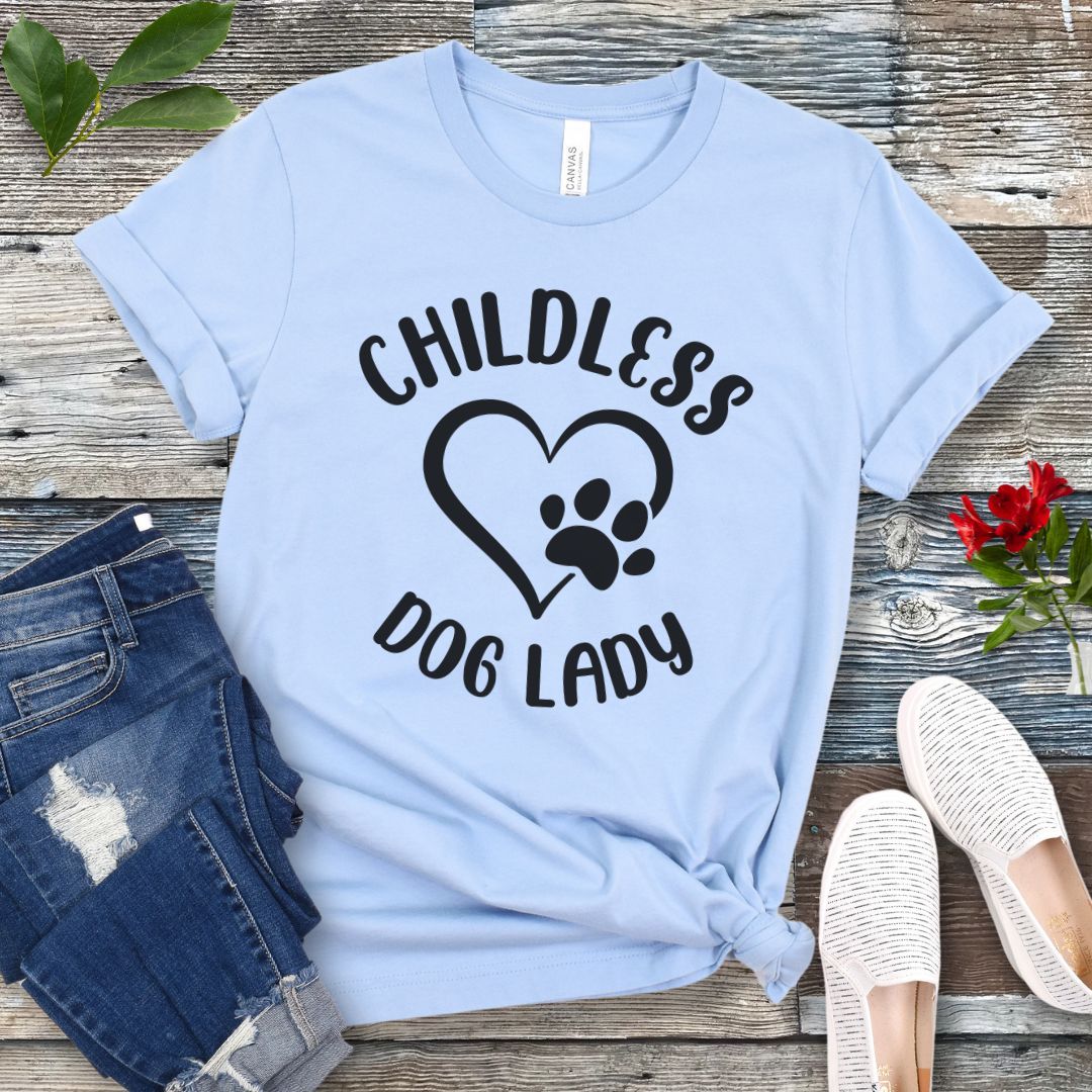 A light blue t-shirt dog dog lovers with the text 'CHILDLESS DOG LADY' in playful lettering and a heart and paw print graphic, displayed on a wooden surface with a pair of jeans and white shoes in the background.