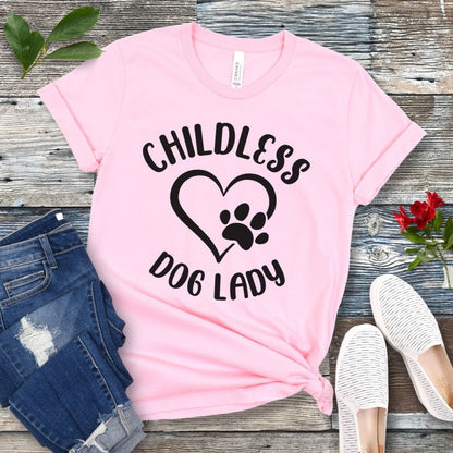 A pink t-shirt dog dog lovers with the text 'CHILDLESS DOG LADY' in playful lettering and a heart and paw print graphic, displayed on a wooden surface with a pair of jeans and white shoes in the background.