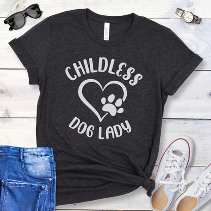 A dark grey t-shirt dog dog lovers with the text 'CHILDLESS DOG LADY' in playful lettering and a heart and paw print graphic, displayed on a wooden surface with a pair of jeans and white shoes in the background.