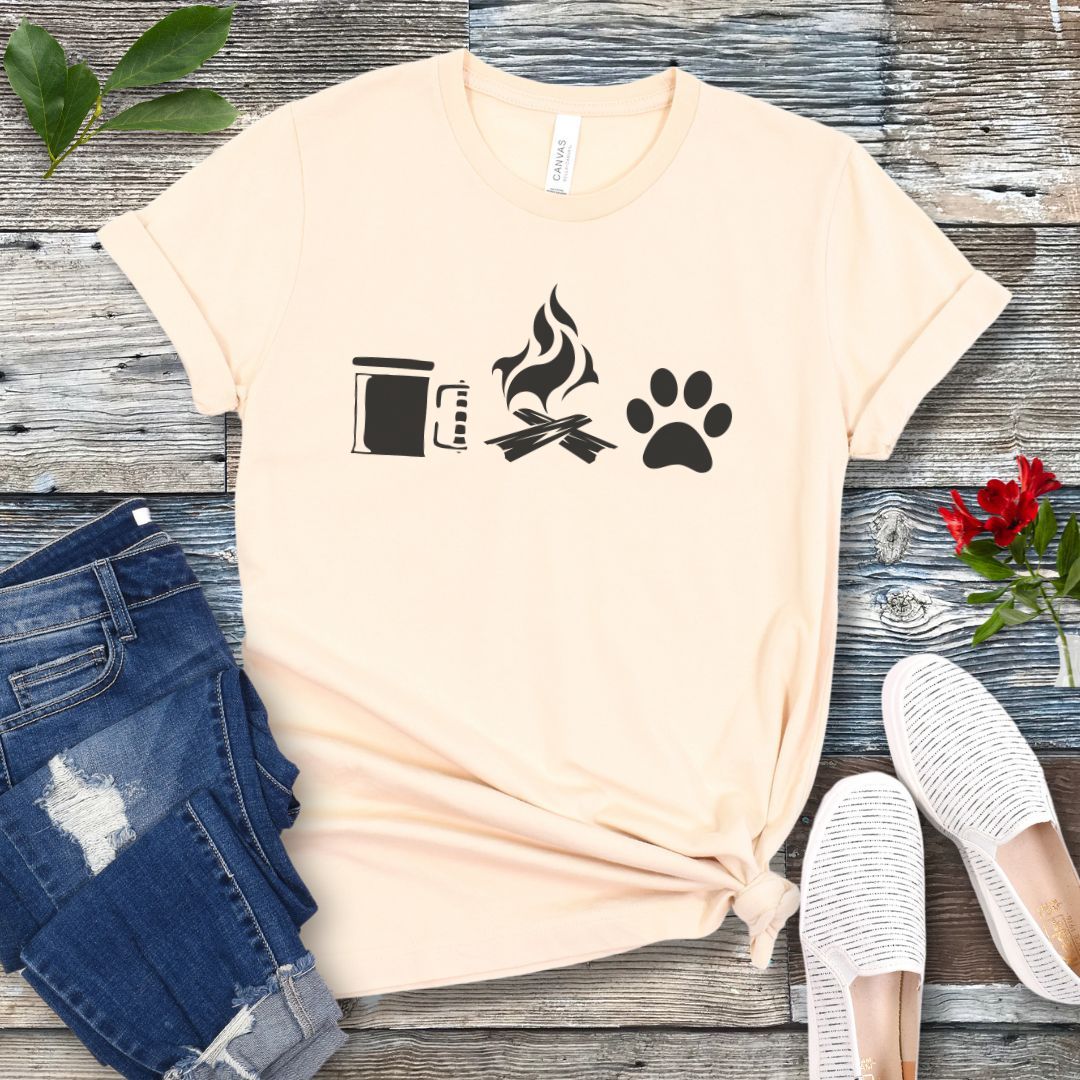 A beige camping and dogs t-shirt with graphics of a coffee cup, campfire, and dog paw print, accompanied by rolled-up blue jeans and white sneakers on a wooden surface.