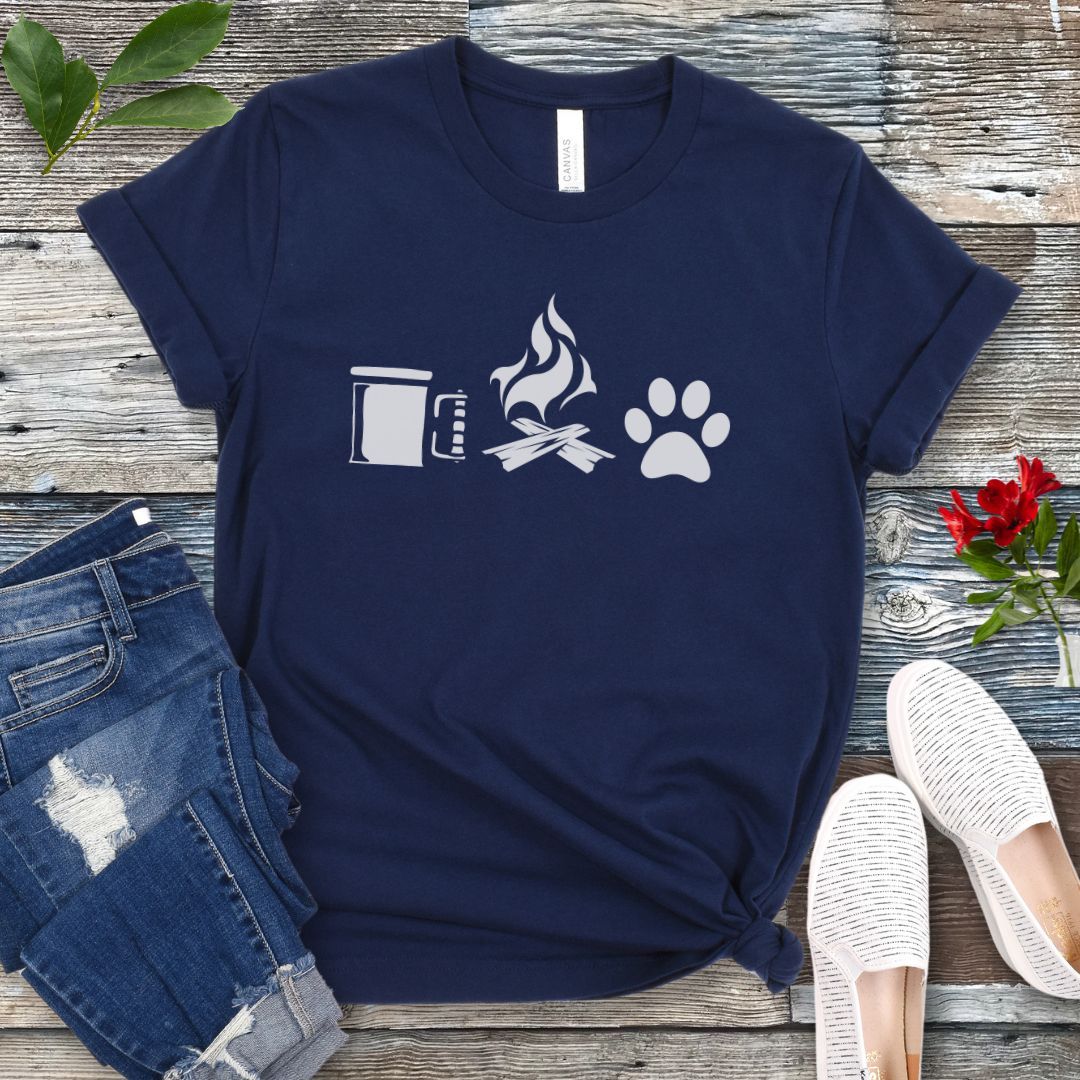 A navy camping and dogs t-shirt with graphics of a coffee cup, campfire, and dog paw print, accompanied by rolled-up blue jeans and white sneakers on a wooden surface.