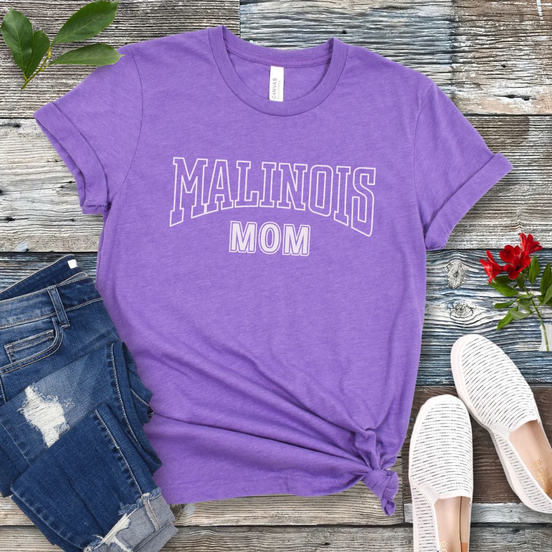A purple Bella Canvas 3001 unisex short sleeve T-shirt with 'MALINOIS MOM' printed in college style lettering, displayed on a wooden surface with denim jeans and white shoes in the background.