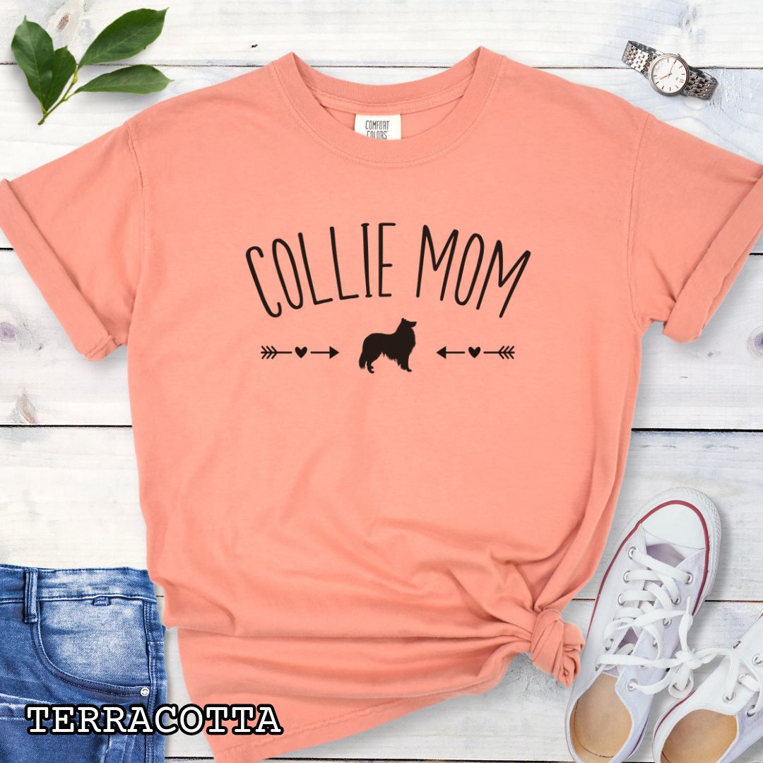 Terracotta Comfort Colors t-shirt with 'Collie Mom' text in playful lettering and Rough Collie dog graphic accented with small hearts and arrows, shown on a wooden surface with sunglasses and jeans.