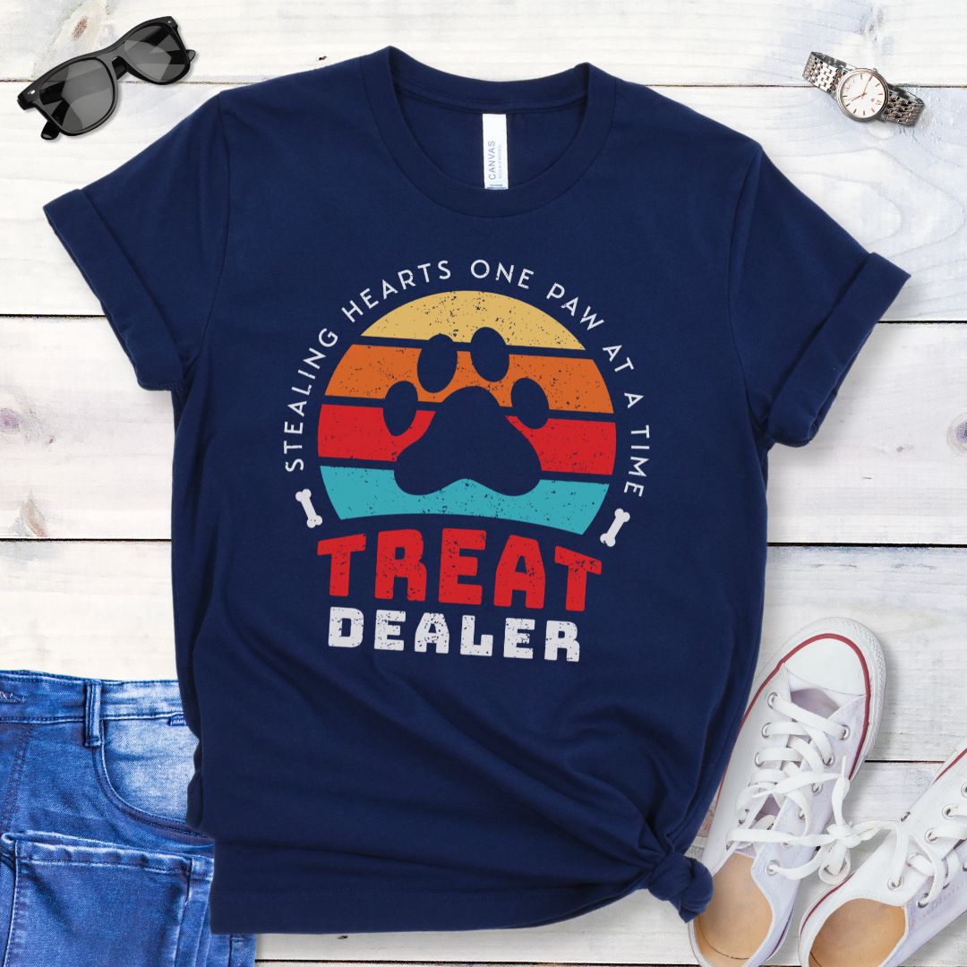 A navy unisex t-shirt for dog lovers with a colorful retro sunset graphic that includes a paw print and the text 'TREAT DEALER HEART STEALER' displayed prominently in a retro style font, laid out flat with a pair of blue jeans and white shoes.
