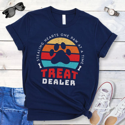 A navy unisex t-shirt for dog lovers with a colorful retro sunset graphic that includes a paw print and the text 'TREAT DEALER HEART STEALER' displayed prominently in a retro style font, laid out flat with a pair of blue jeans and white shoes.