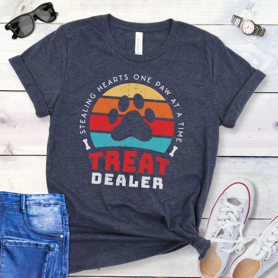 A heather navy unisex t-shirt for dog lovers with a colorful retro sunset graphic that includes a paw print and the text 'TREAT DEALER HEART STEALER' displayed prominently in a retro style font, laid out flat with a pair of blue jeans and white shoes.