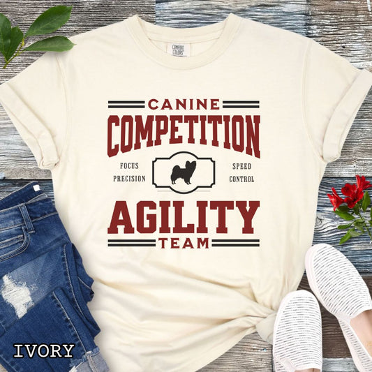 Beige Comfort Colors dog agility t-shirt with 'Canine Competition Agility Team' design and custom breed silhouette, presented on a wooden surface with denim shorts and a flower.