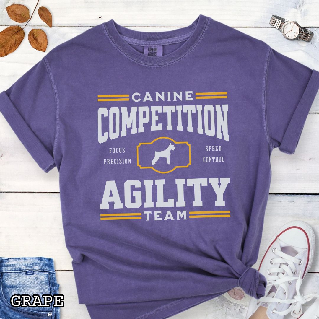 Purple Comfort Colors dog agility t-shirt with 'Canine Competition Agility Team' design and custom breed silhouette, presented on a wooden surface with denim shorts and a flower.