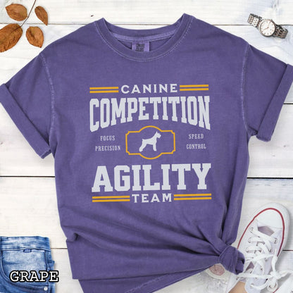Purple Comfort Colors dog agility t-shirt with 'Canine Competition Agility Team' design and custom breed silhouette, presented on a wooden surface with denim shorts and a flower.