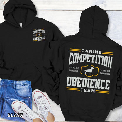 Black dog obedience back print hoodie with 'Canine Competition Obedience Team' text and custom breed silhouette, presented on a wooden surface with denim jeans and a flower.