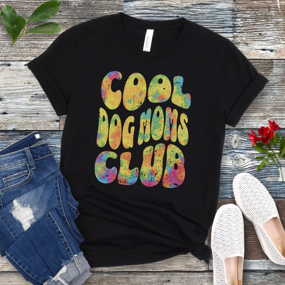A black dog lover t-shirt with the text 'COOL DOG MOMS CLUB' printed in a colorful, groovy font, displayed on a wooden surface with a pair of jeans and white shoes in the background.
