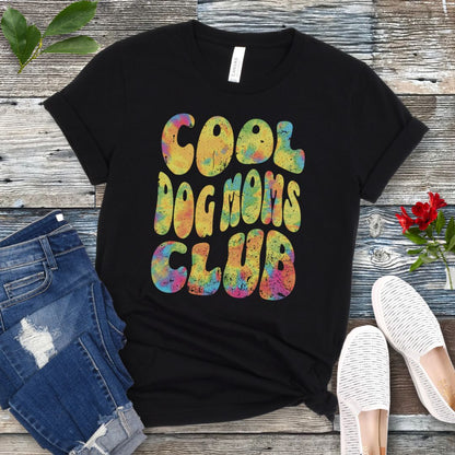 A black dog lover t-shirt with the text 'COOL DOG MOMS CLUB' printed in a colorful, groovy font, displayed on a wooden surface with a pair of jeans and white shoes in the background.
