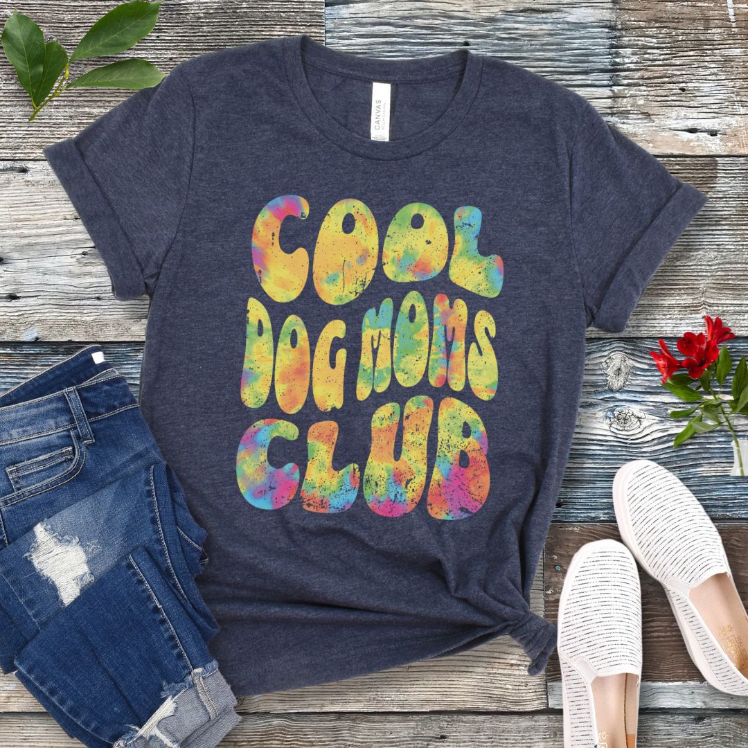 A heather navy dog lover t-shirt with the text 'COOL DOG MOMS CLUB' printed in a colorful, groovy font, displayed on a wooden surface with a pair of jeans and white shoes in the background.