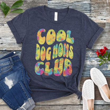 A heather navy dog lover t-shirt with the text 'COOL DOG MOMS CLUB' printed in a colorful, groovy font, displayed on a wooden surface with a pair of jeans and white shoes in the background.