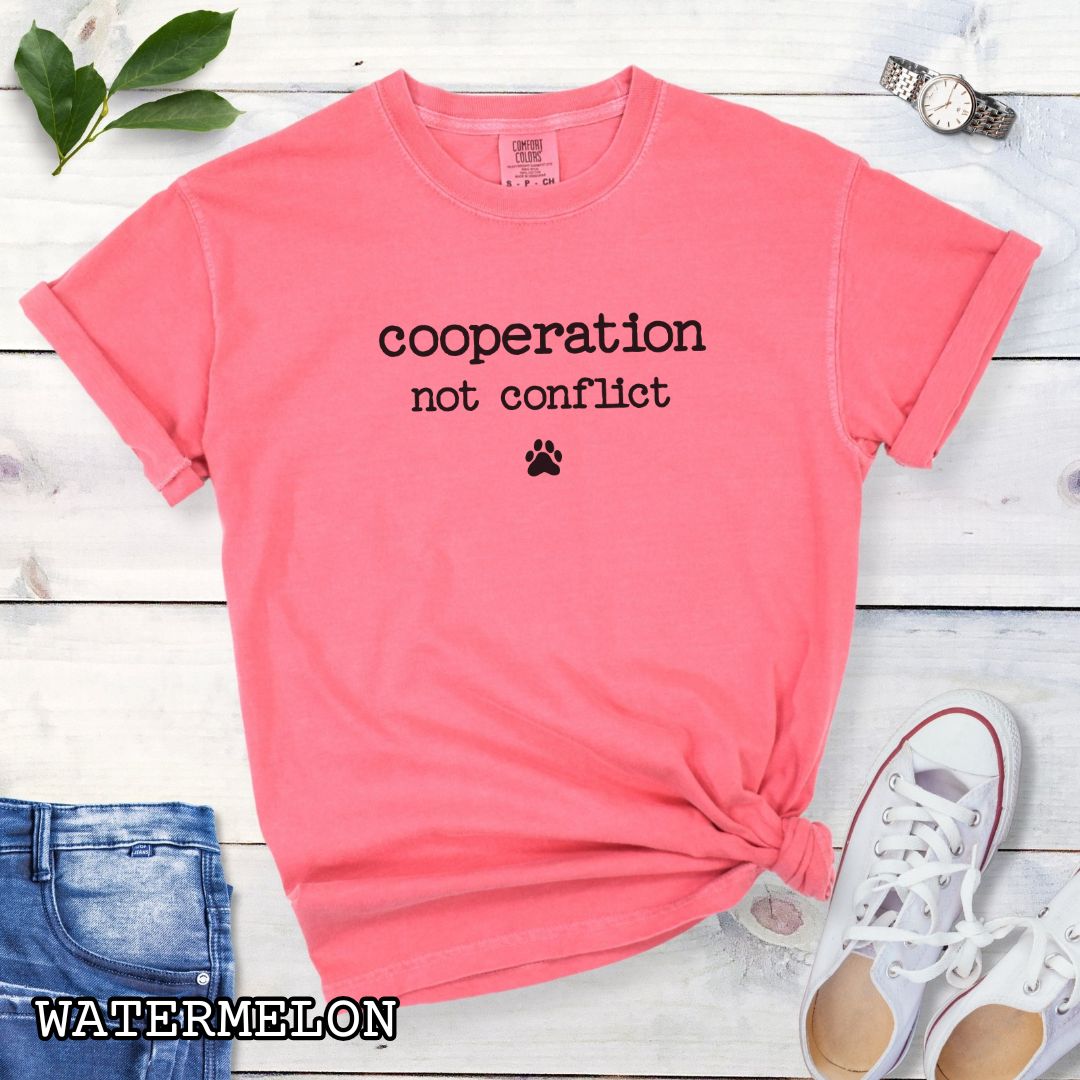 Pink (Watermelon) Comfort Colors dog training t-shirt with 'cooperation not conflict' text on a wooden surface with shoes and jeans.