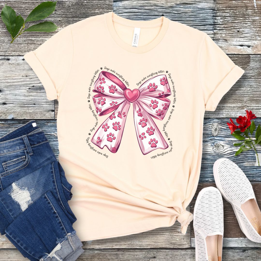 A beige, coquette-style dog lover t-shirt with a bow design featuring a bow design filled with pink paw prints, surrounded by the script text 'dogs make everything better', displayed flat with a pair of blue denim shorts and white shoes on a wooden surface.