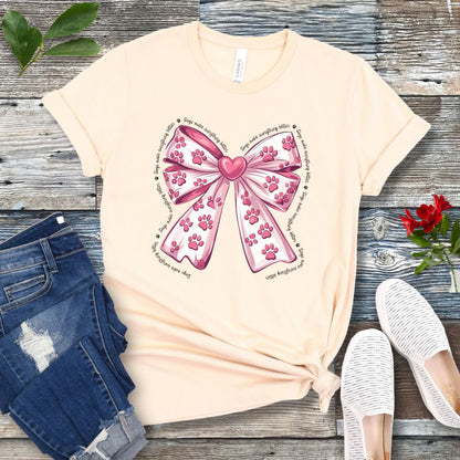 A beige, coquette-style dog lover t-shirt with a bow design featuring a bow design filled with pink paw prints, surrounded by the script text 'dogs make everything better', displayed flat with a pair of blue denim shorts and white shoes on a wooden surface.