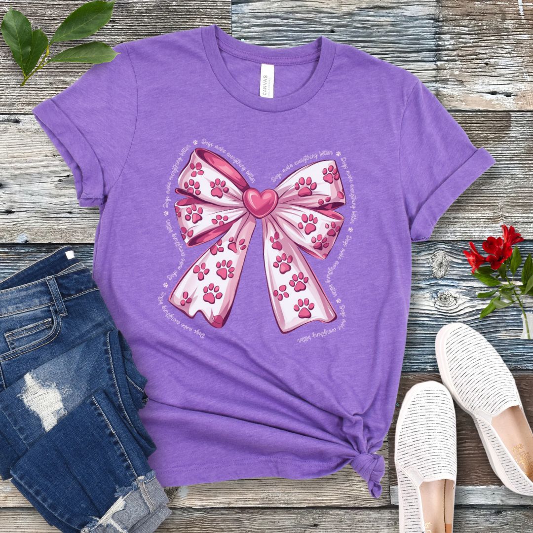 A purple, coquette-style dog lover t-shirt with a bow design featuring a bow design filled with pink paw prints, surrounded by the script text 'dogs make everything better', displayed flat with a pair of blue denim shorts and white shoes on a wooden surface.
