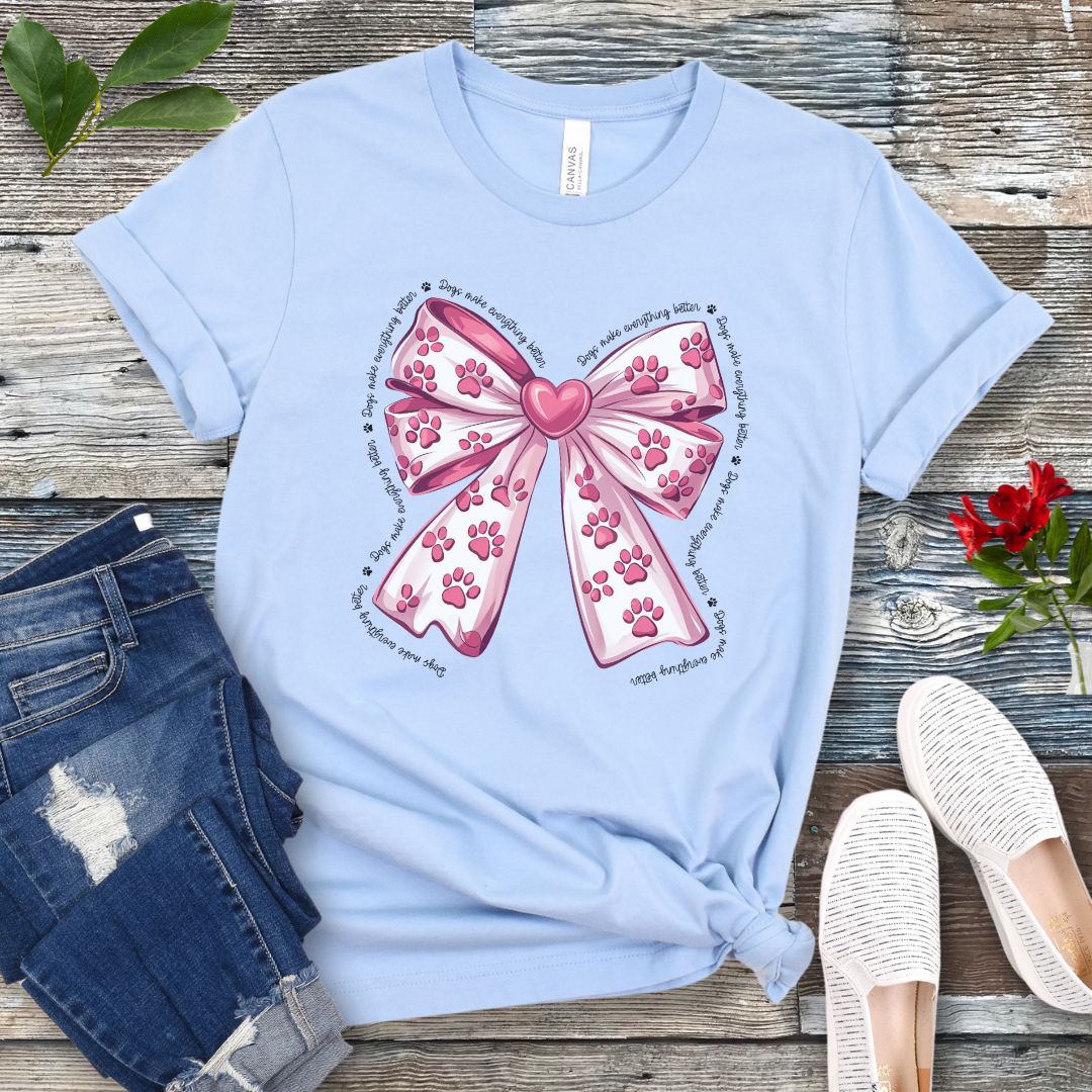 A light blue, coquette-style dog lover t-shirt with a bow design featuring a bow design filled with pink paw prints, surrounded by the script text 'dogs make everything better', displayed flat with a pair of blue denim shorts and white shoes on a wooden surface.